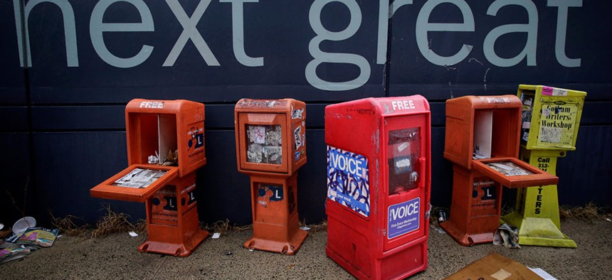 Plastic newspaper racks for The Village Voice and other newspapers stand along a Manhattan sidewalk.