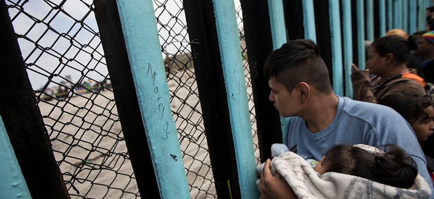 An asylum-seeking migrant from Central America holds a child while peering through the border wall in Tijuana, Mexico.