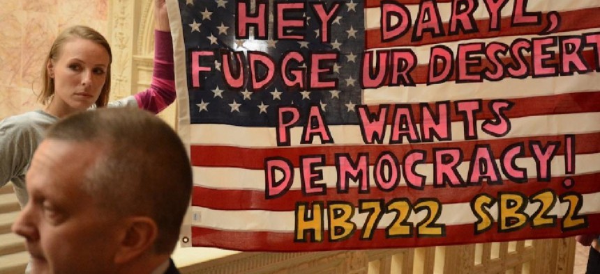 A member of Fair Districts PA holds up a banner urging House State Government Committee Chairman Daryl Metcalfe (foreground) to vote House Bill 722, which would establish an independent commission for redistricting, out of committee.