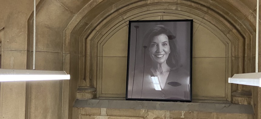 Several reporters gathered as Office of General Services staffers placed Gov. Kathy Hochul’s portrait above the Capitol Pressroom studio today.