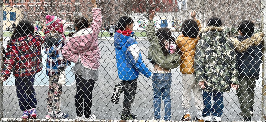 A row of students ages 5-7 raising their hands while standing along a chain-link fence on a playground during recess at P.S. 111, a public school in Hell's Kitchen, New York City.