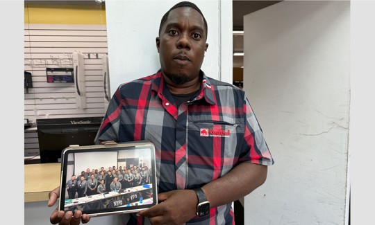 Marquis Anderson at his job working in a beverage distribuion warehouse holds a photo of himself as part of the October 2022 NYPD Academy Class. 