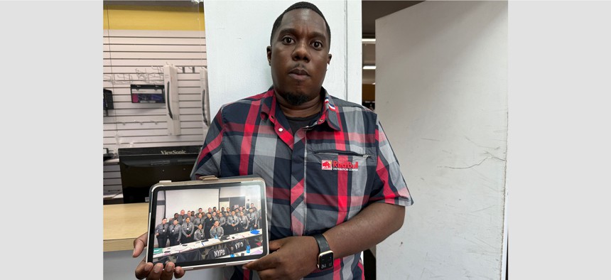 Marquis Anderson at his job working in a beverage distribuion warehouse holds a photo of himself as part of the October 2022 NYPD Academy Class. 